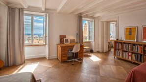 A spacious room with a wooden desk, a bookshelf, and large windows offering a view of buildings and a tower. - Le Saint-Jean (Bastia)