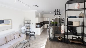 Uma sala de estar moderna com um sofá, mesa de centro e uma estante. Uma cozinha é visível ao fundo.