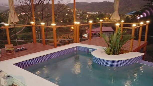 Ein Swimmingpool mit einer bepflasterten Kante, umgeben von einer Holzterrasse und einer Pergola mit Lichterketten.