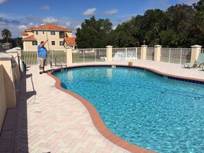Une piscine entourée d’une terrasse en briques et d’une clôture blanche, avec, en arrière-plan, un bâtiment dont le toit est recouvert de tuiles rouges.