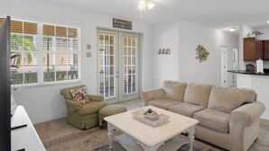 En stue med en beige sofa, et hvidt sofabord og en grøn lænestol. Der er to vinduer med persienner, en loftsvifte og et køkken, der kan ses gennem en åben dør.