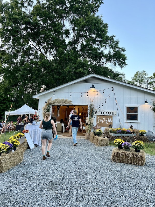 Un bâtiment rustique, agrémenté d’une pancarte « Bienvenue », avec un sol en gravier et des espaces de repos extérieurs comprenant des bottes de foin et des fleurs.