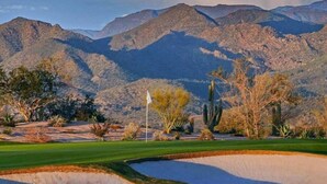 Un campo de golf con una bandera, cactus y montañas al fondo.