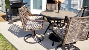 Een terras buiten met rieten meubilair en een barbecue, voor een lichtblauwe gevel met witte accenten.
