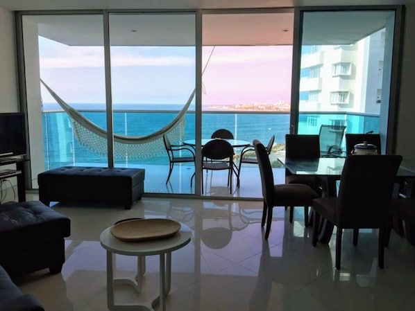A spacious living area with a large sliding glass door leading to a balcony with a view of the sea and a hammock.