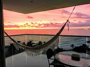 A balcony with a glass table and chairs, a hammock, and a view of the ocean at sunset. - Sea Terraces Luxury Suites (Cartagena)