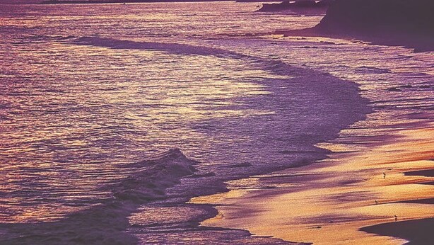 Atardecer sobre una playa con olas y un muelle a lo lejos.