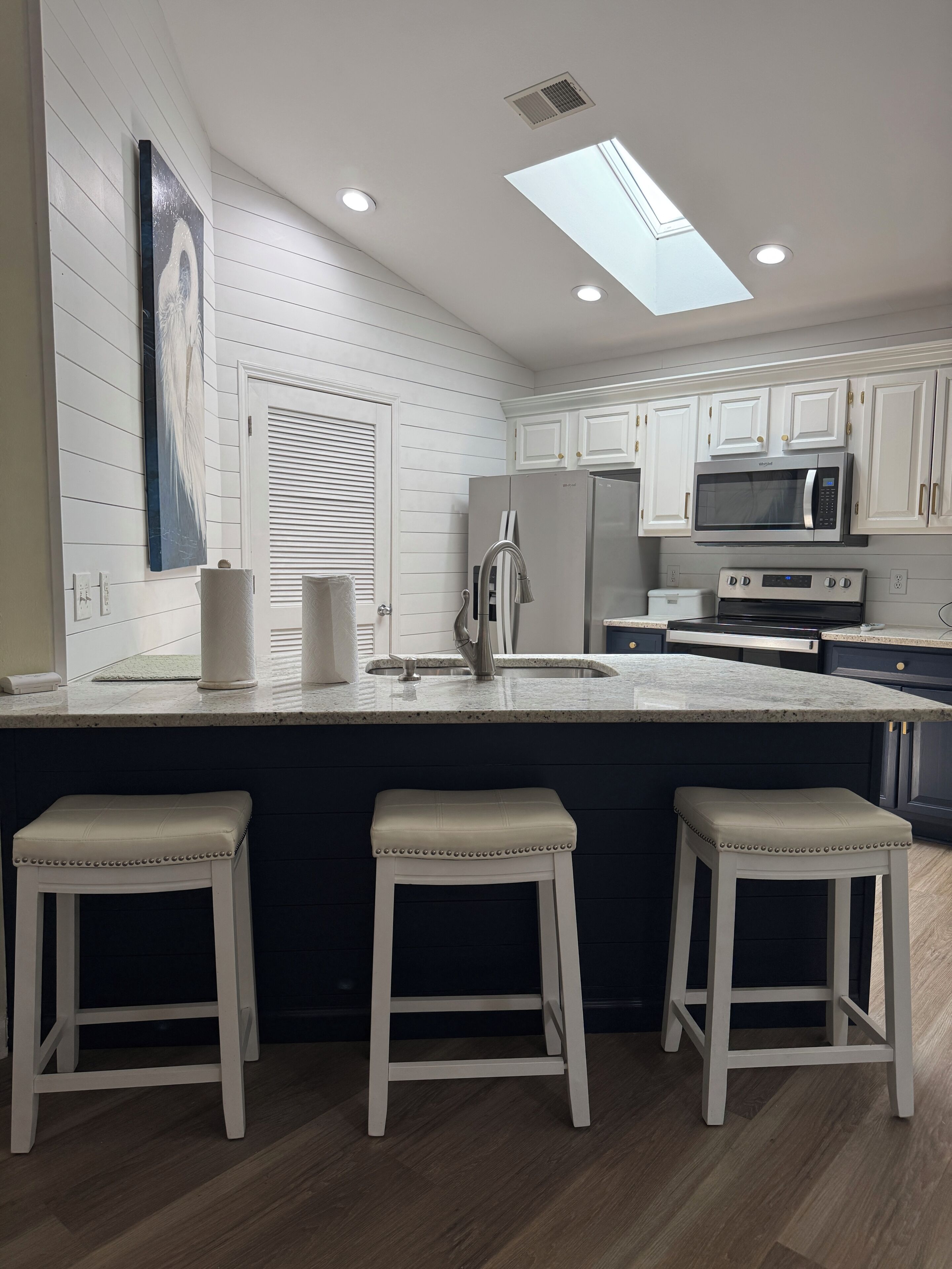 A kitchen with white cabinets, a stainless steel refrigerator, a microwave, a sink, and three bar stools.