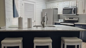 Cocina con gabinetes blancos, refrigerador de acero inoxidable, microondas, fregadero y tres taburetes de bar.