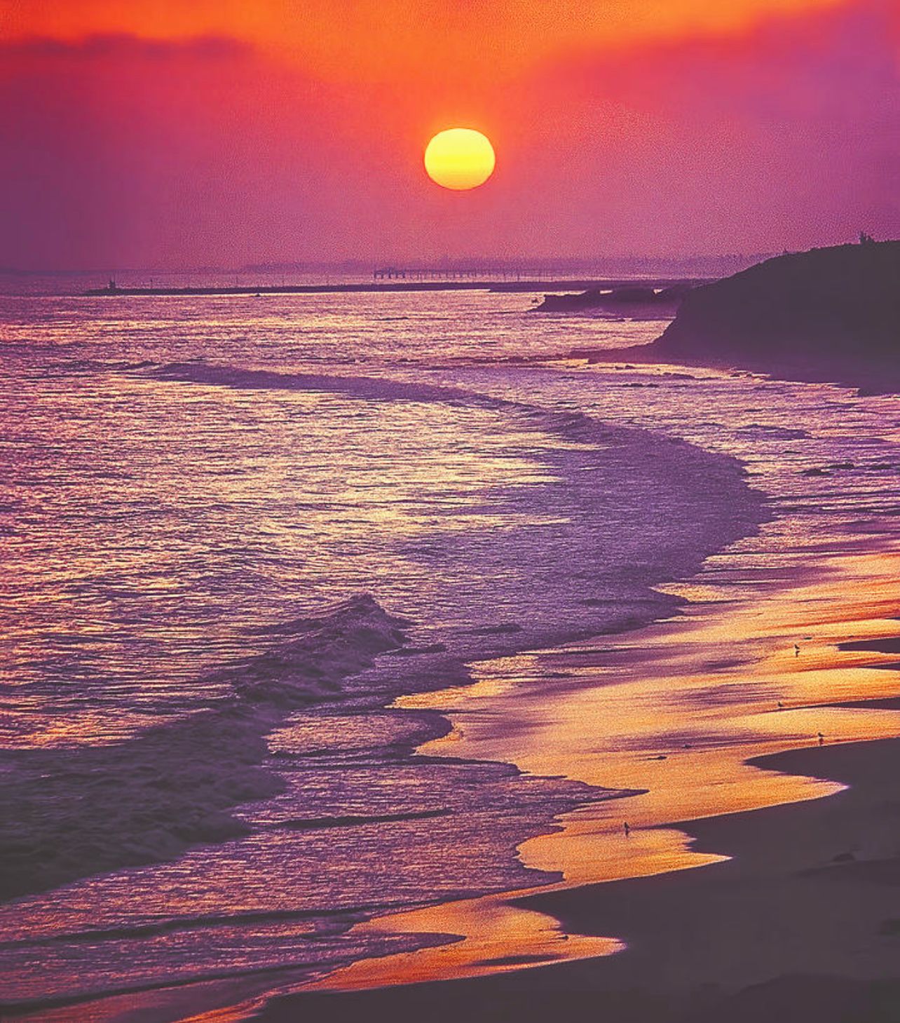A sunset over a beach with waves and a pier in the distance.