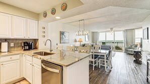 A spacious kitchen with white cabinets, a built-in dishwasher, and a large island. There is a dining area with a table and chairs, and a balcony with a view. - Oceanfront Beach Condo w/ Stunning Views! (North Myrtle Beach)