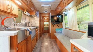 Cocina compacta para autocaravana con armarios de madera, un lavamanos, estufa y un televisor en un asiento junto a la ventana.