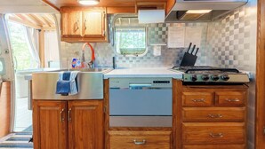 Cocina compacta para casa rodante con armarios de madera, lavavajillas, estufa y ventana con vista a árboles.