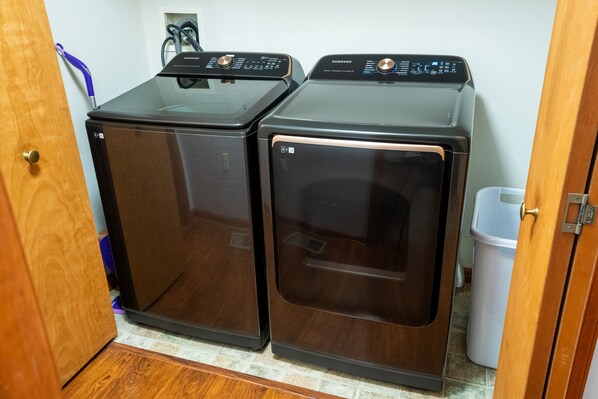 Two front-loading washing machines in a laundry room with a door on the left and a trash can on the right.