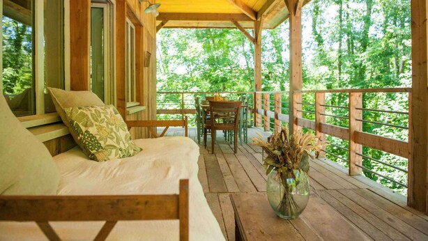 A wooden deck with a sofa and chairs, a table with a vase of dried flowers, and a view of a forest.