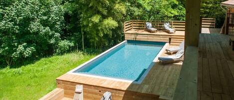 Une piscine rectangulaire entourée d’une végétation luxuriante, avec une terrasse en bois comprenant des sièges et des chaises longues.