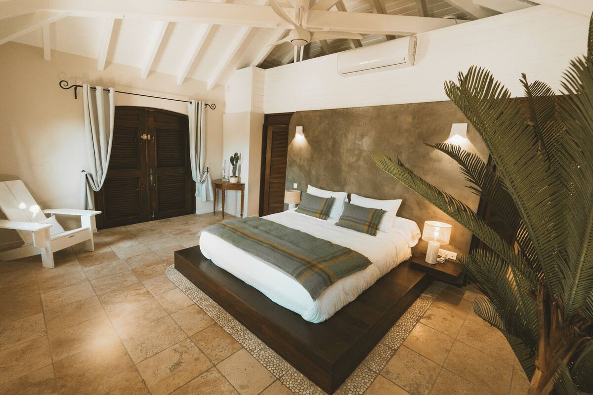 A bedroom with a large bed, a chair, a nightstand, and a potted plant.