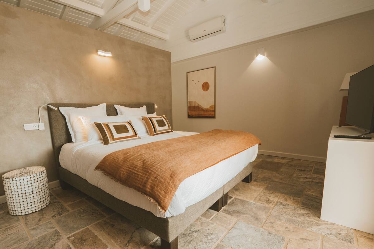 A bedroom with a large bed, a round side table, a wall-mounted air conditioner, and a painting on the wall.