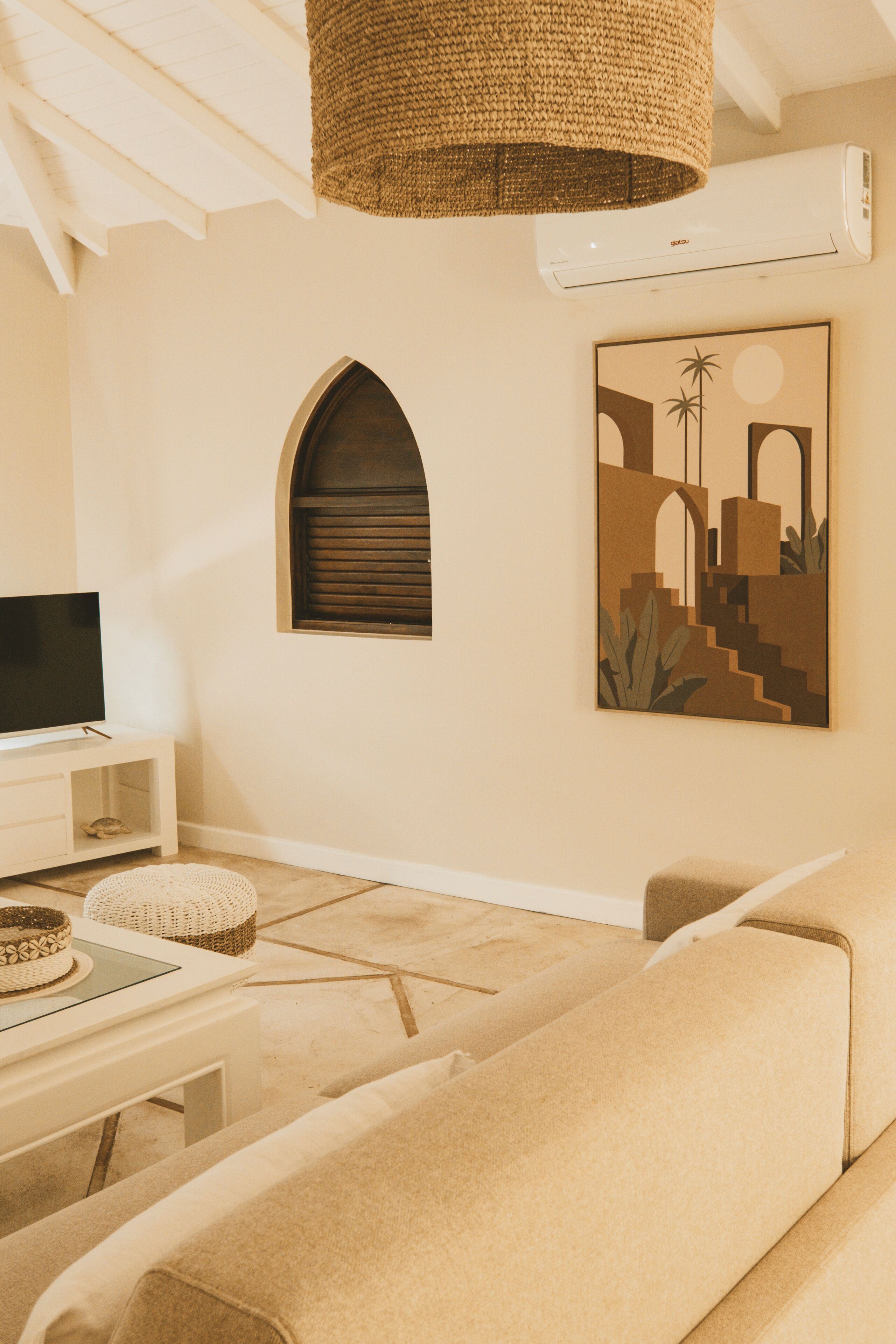 A living room with a beige sofa, a white entertainment unit, a woven hanging light fixture, and a wall-mounted air conditioner.