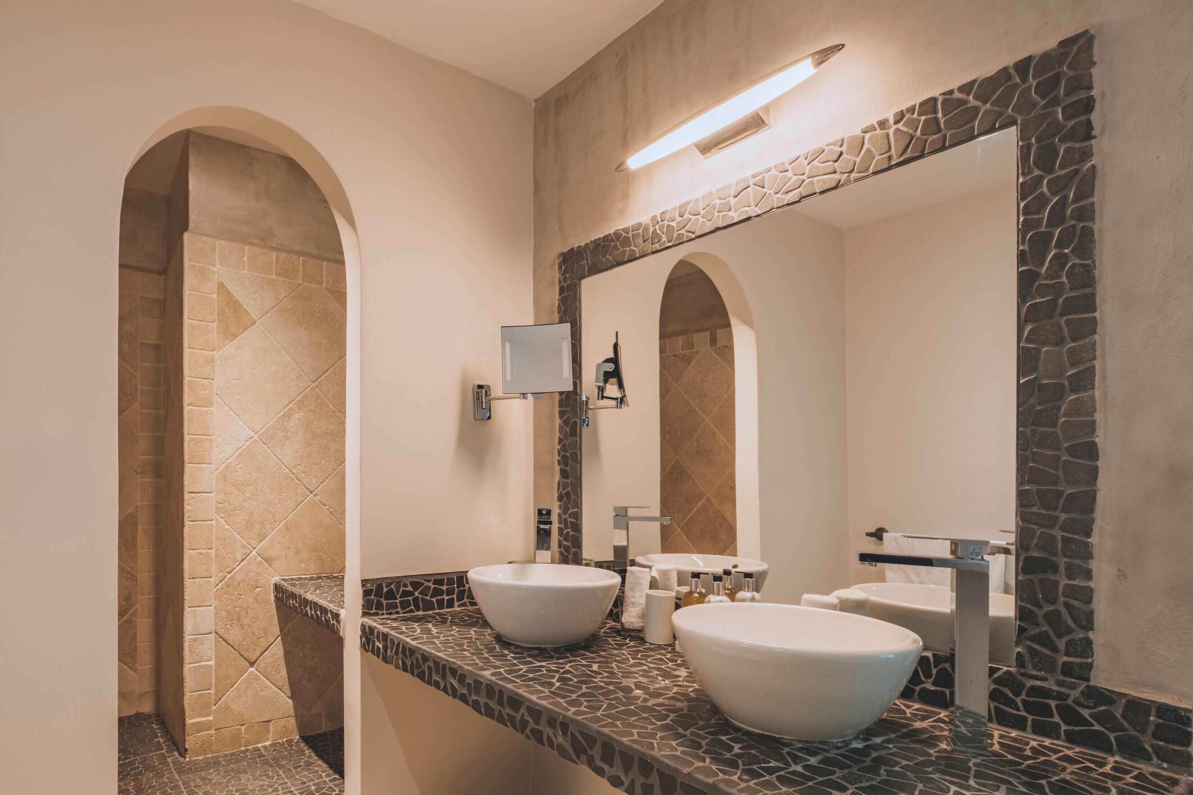 A modern bathroom with two large white sinks, a stone accent wall, and a large mirror.