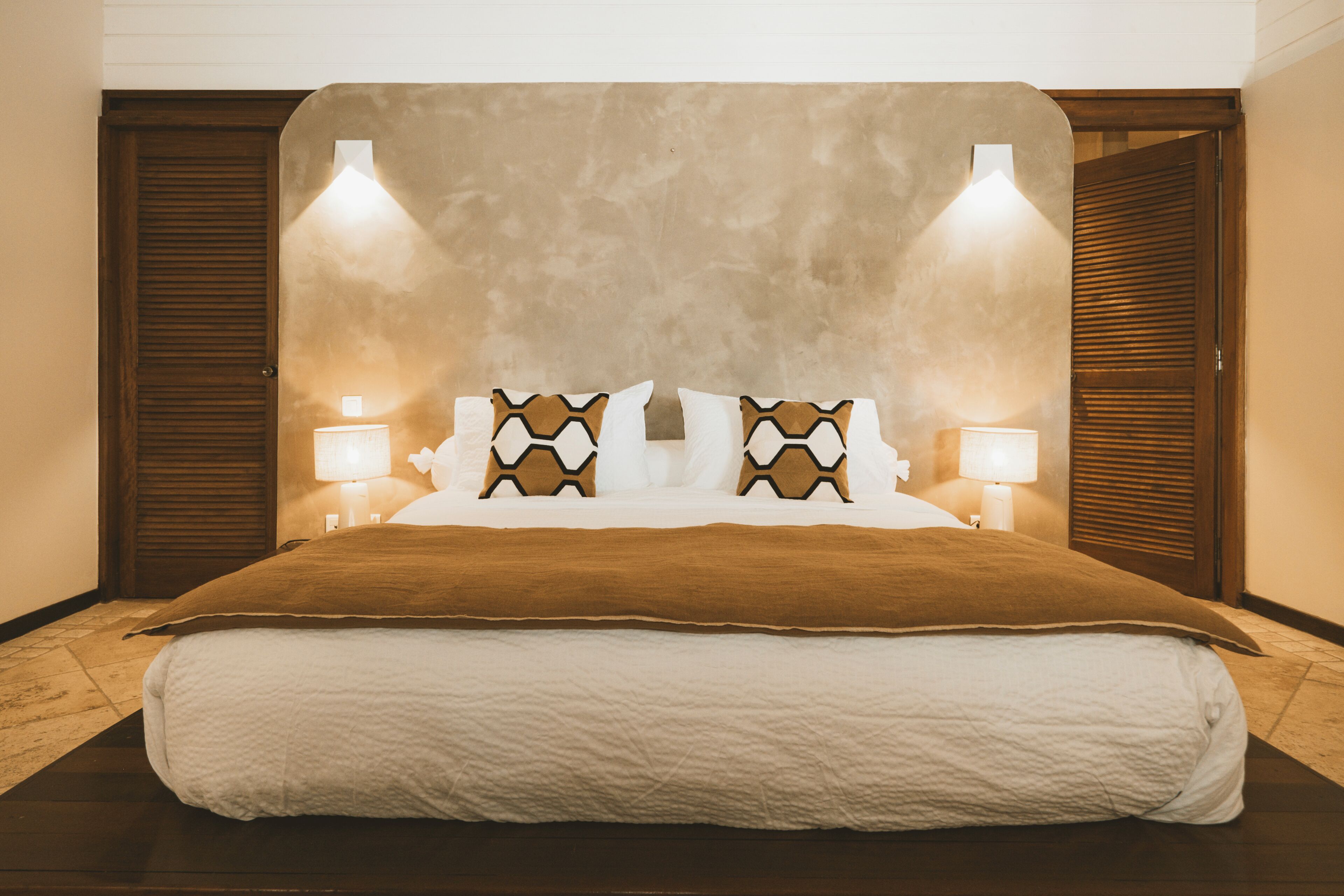 A large bed with a brown headboard, white bedding, and decorative pillows. There are two bedside lamps on either side of the bed.
