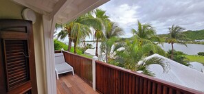 Un balcon en bois avec un fauteuil de jardin blanc, donnant sur un lac bordé de palmiers et un paysage vallonné.