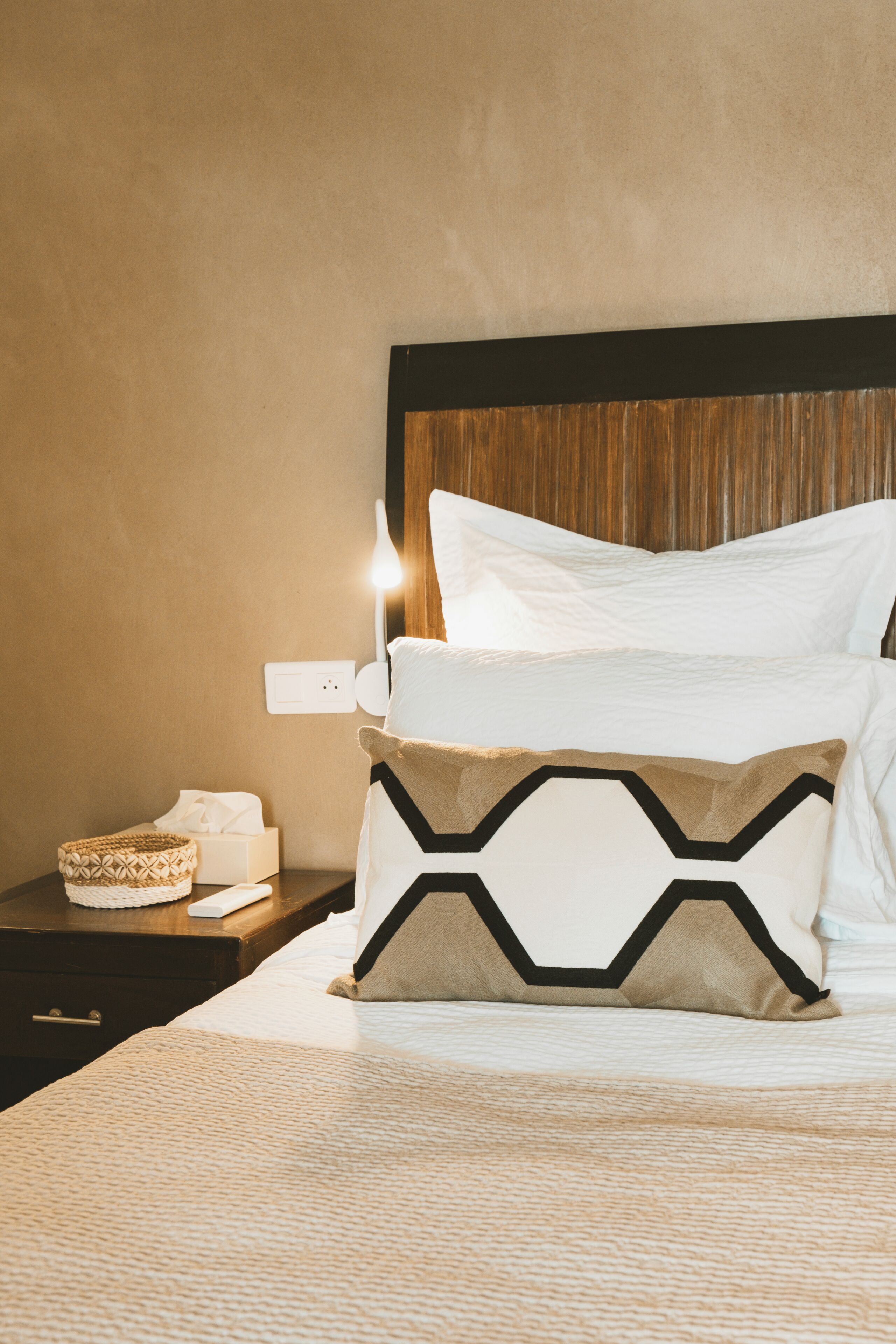Un lit bien fait, avec des oreillers blancs et à motifs, une lampe de chevet et un petit panier posé sur une table de chevet en bois.