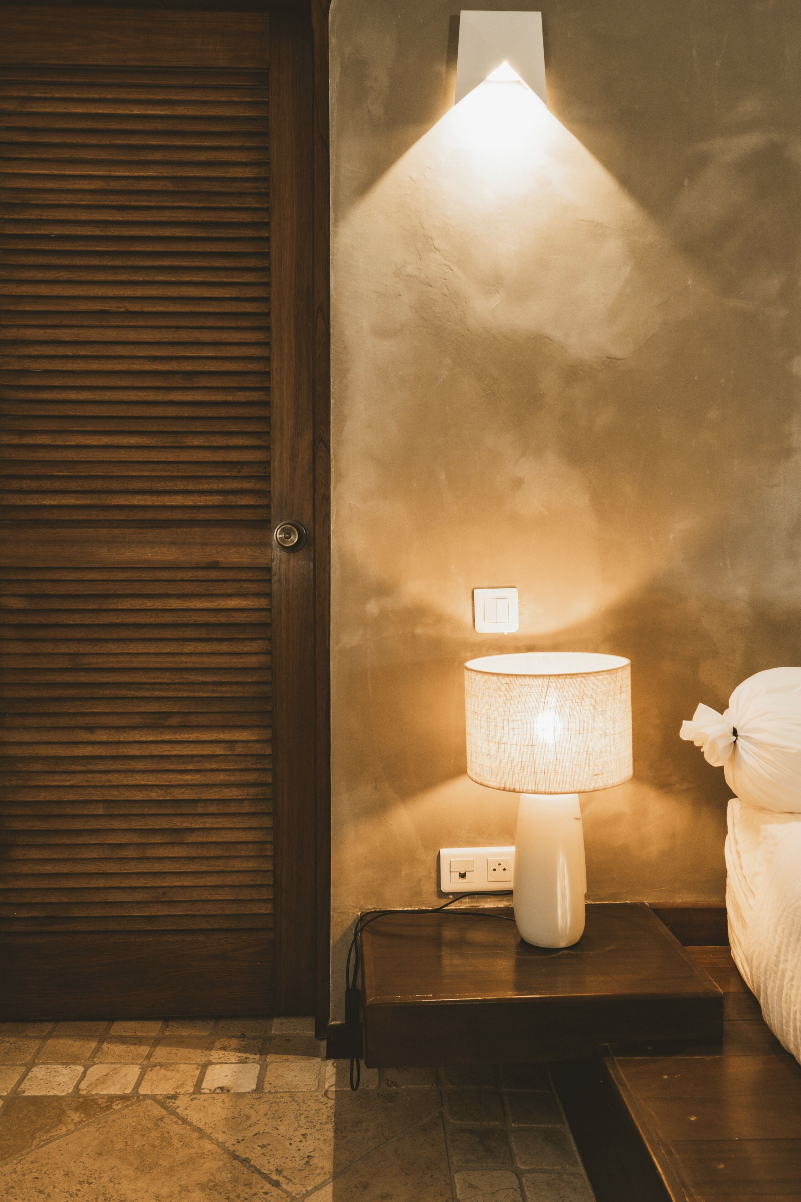 A room with a wooden door, a nightstand with a lamp, and a bed with a white pillow.