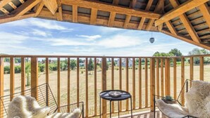 A wooden deck with a view of a grassy field, a hanging light fixture, and two chairs with fluffy cushions.