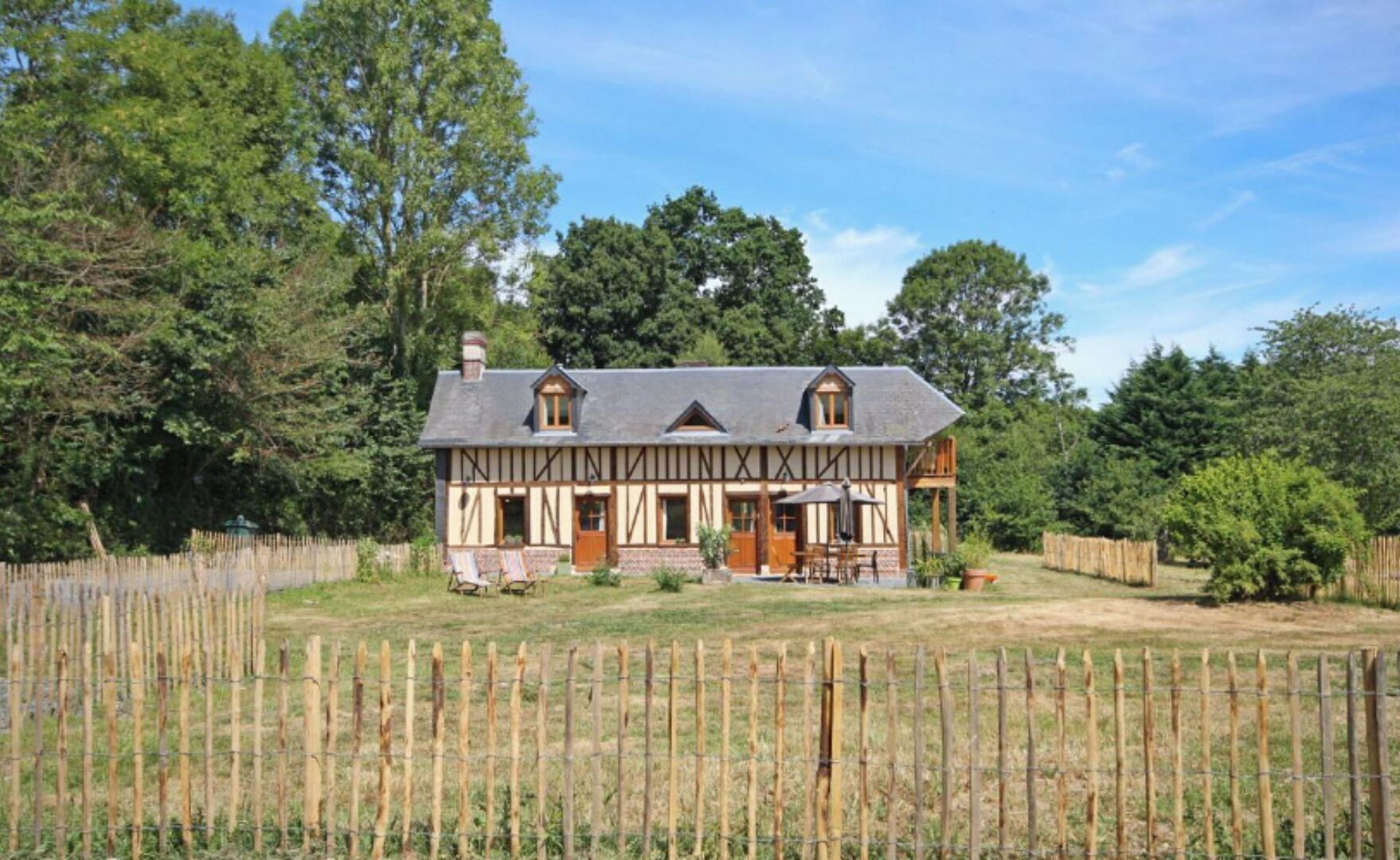 Une maison traditionnelle à colombages, dotée d’un grand porche à l’avant, entourée d’une clôture en bois et d’une végétation luxuriante.