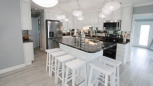 Una cucina moderna con mobili bianchi, una grande isola con sgabelli da bar, elettrodomestici in acciaio inossidabile e lampadari di cristallo.