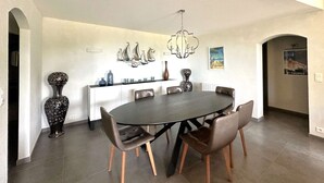 Un comedor con una mesa redonda, seis sillas, una araña, jarrones decorativos y una obra de arte colgada en la pared.
