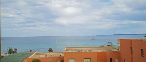 A balcony with a view of the sea and distant land, featuring a building with a flat roof and a small sailboat in the water.