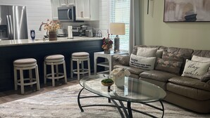 Una cocina moderna con armarios blancos, una isla central con taburetes, una zona de comedor con mesa de vidrio y una sala de estar con sofá y cojines.