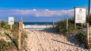 Een zandpad langs het strand dat naar de oceaan leidt, met borden die toegang tot het openbare strand en veiligheidsinformatie aangeven.