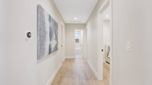 A hallway with light-colored walls, wooden flooring, and a ceiling light. There are doors on the right, and a large abstract painting on the left. - Upscale Family Home · 5 miles to Disneyland (Fullerton)