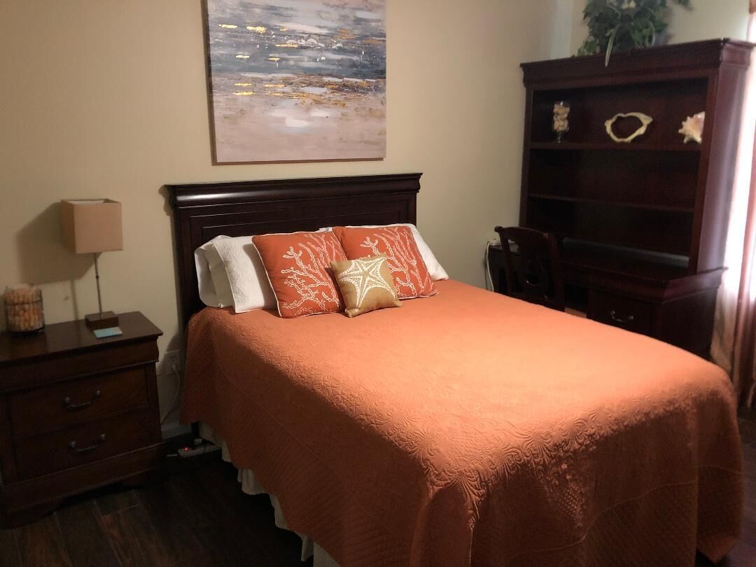 A neatly made bed with a brown bedspread and two decorative pillows, a wooden nightstand with a lamp, a small cabinet, and a large painting on the wall.