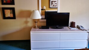 Habitación de hotel con tocador blanco, televisor de pantalla plana, lámpara, teléfono y dos cuadros en la pared.