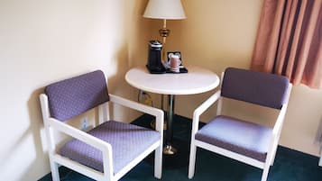 A small room with two white chairs with patterned cushions, a round white table, a lamp, a coffee maker, and a brown curtain.
