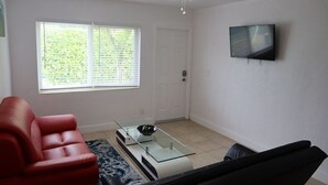 A living room with a red sofa, a glass coffee table, a ceiling fan, and a TV mounted on the wall.