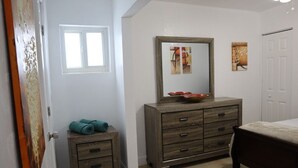 A bedroom with a wooden dresser, a bed, a small table with towels, and a window.