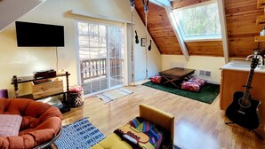 Ein geräumiger Loft mit hohen Decken, Holzbalken, einer Glastür, die nach draußen führt, einer gemütlichen Sitzecke mit Sofa und Sesseln, einem kleinen Tisch und einer Gitarre.