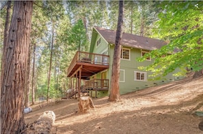 Une cabane à deux étages dotée d’un balcon, entourée de grands arbres et bordée d’un sentier de terre.