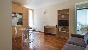 Un salon moderne avec un plancher en bois, une table à manger en verre, un canapé, un téléviseur et un coin cuisine doté de meubles en bois et d’une fenêtre.