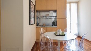 Une cuisine compacte avec des meubles en bois, un plan de travail blanc et une table de salle à manger ronde, assortie de chaises transparentes.