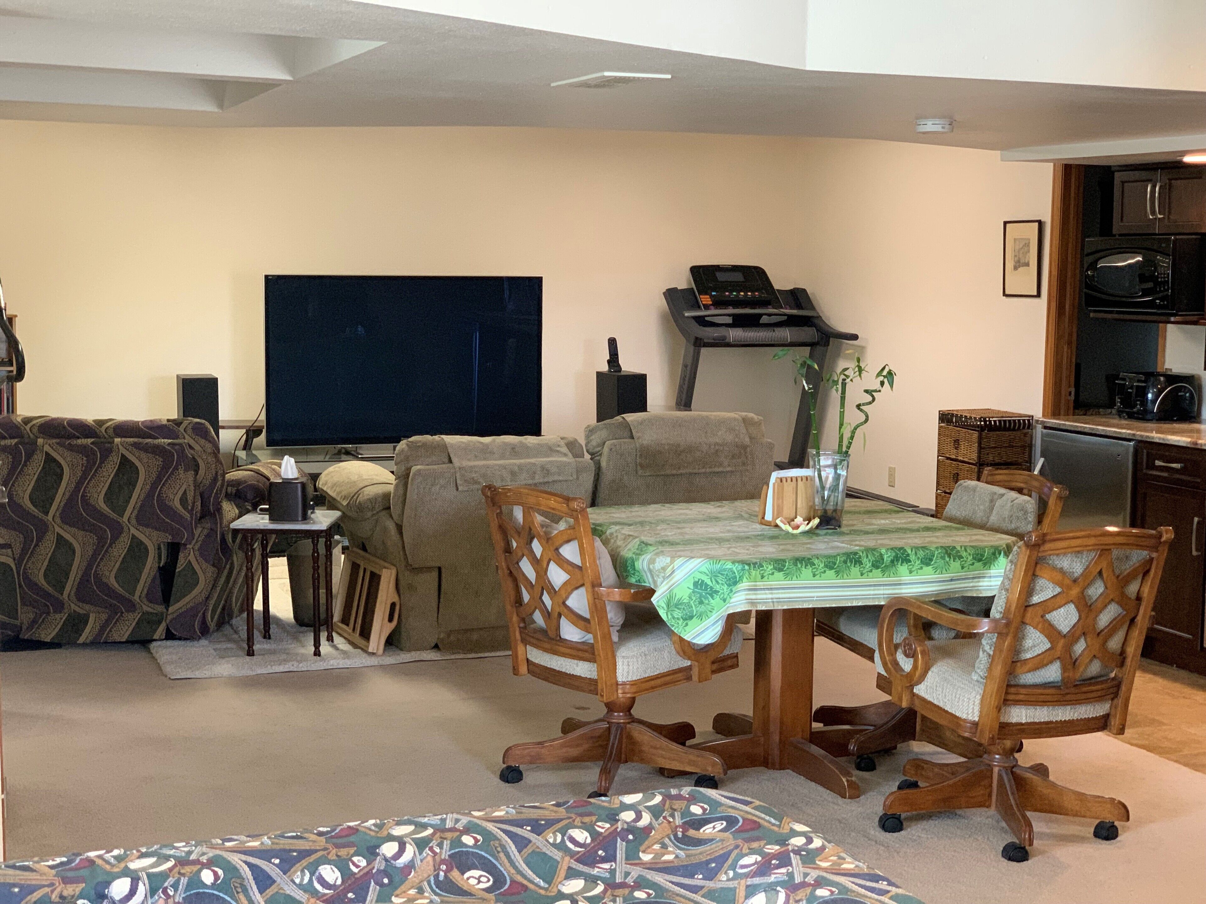 A living room with a sofa, a dining table with chairs, a treadmill, a TV, and a kitchen area with appliances.