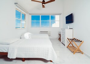 A bright, minimalist bedroom with two beds, a ceiling fan, a large window, a TV, and a wooden drying rack. - #20 Luxury True Oceanfront Designer Penthouse (Isabela)