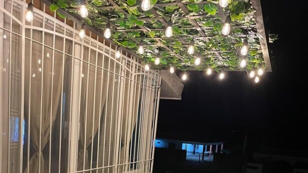 Zona d'estar a l'aire lliure amb mobles de vimetes i taula de vidre, sota una pérgola amb llums de cadena.