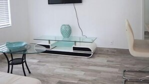 Ein Zimmer mit Flachbildfernseher an der Wand, einem Glastisch, einem weißen Sofa und einem Deckenventilator.