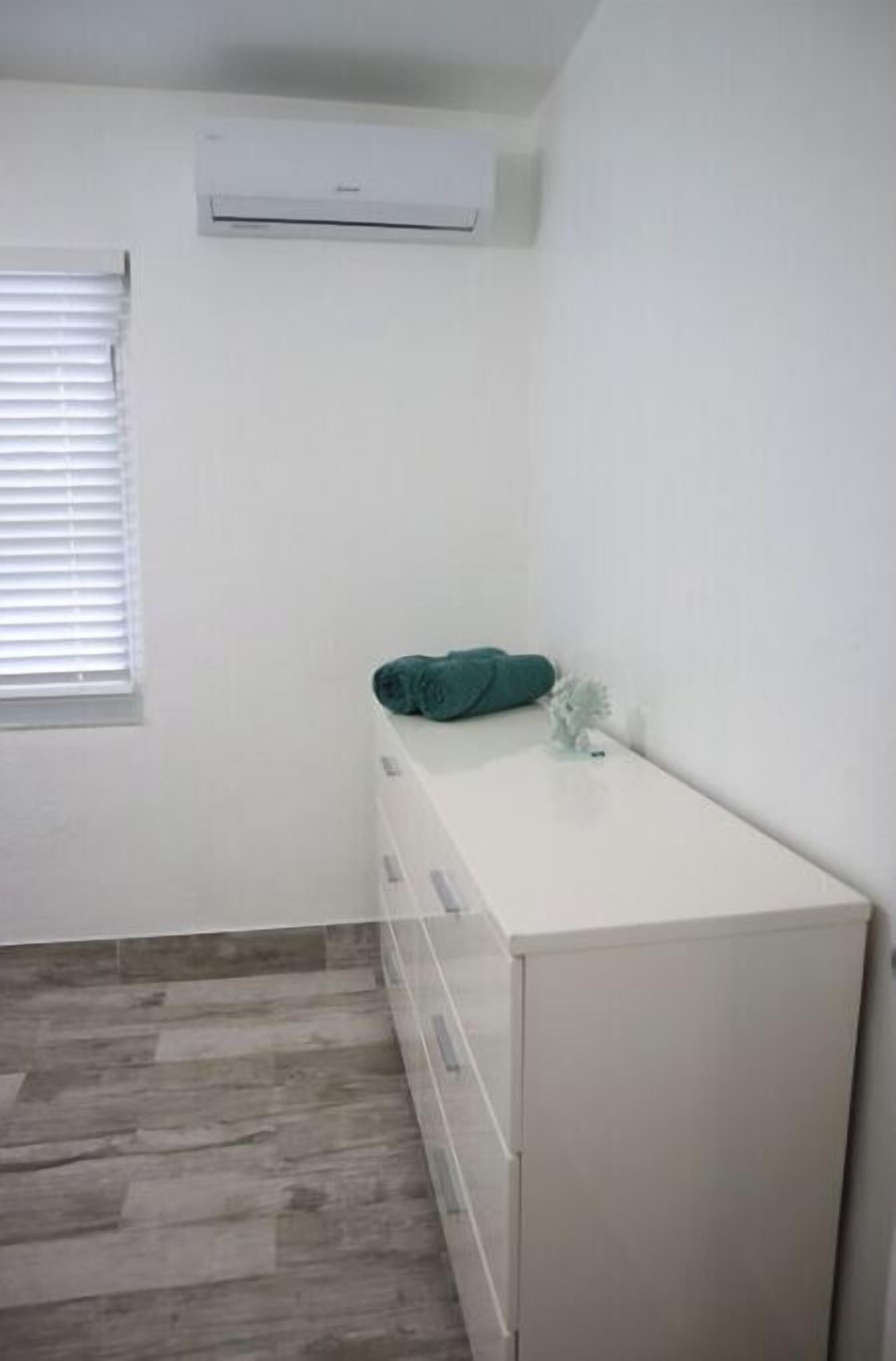Une petite pièce propre, avec une commode blanche, une fenêtre équipée de stores et un radiateur blanc.